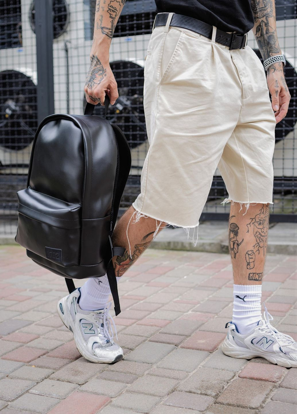Новини Хмельницького - фото з Як обрати рюкзак чоловічий онлайн і не пошкодувати про покупку (новини компаній) Чоловік із татуюваннями у бежевих шортах та білих кросівках тримає чорний міський рюкзак